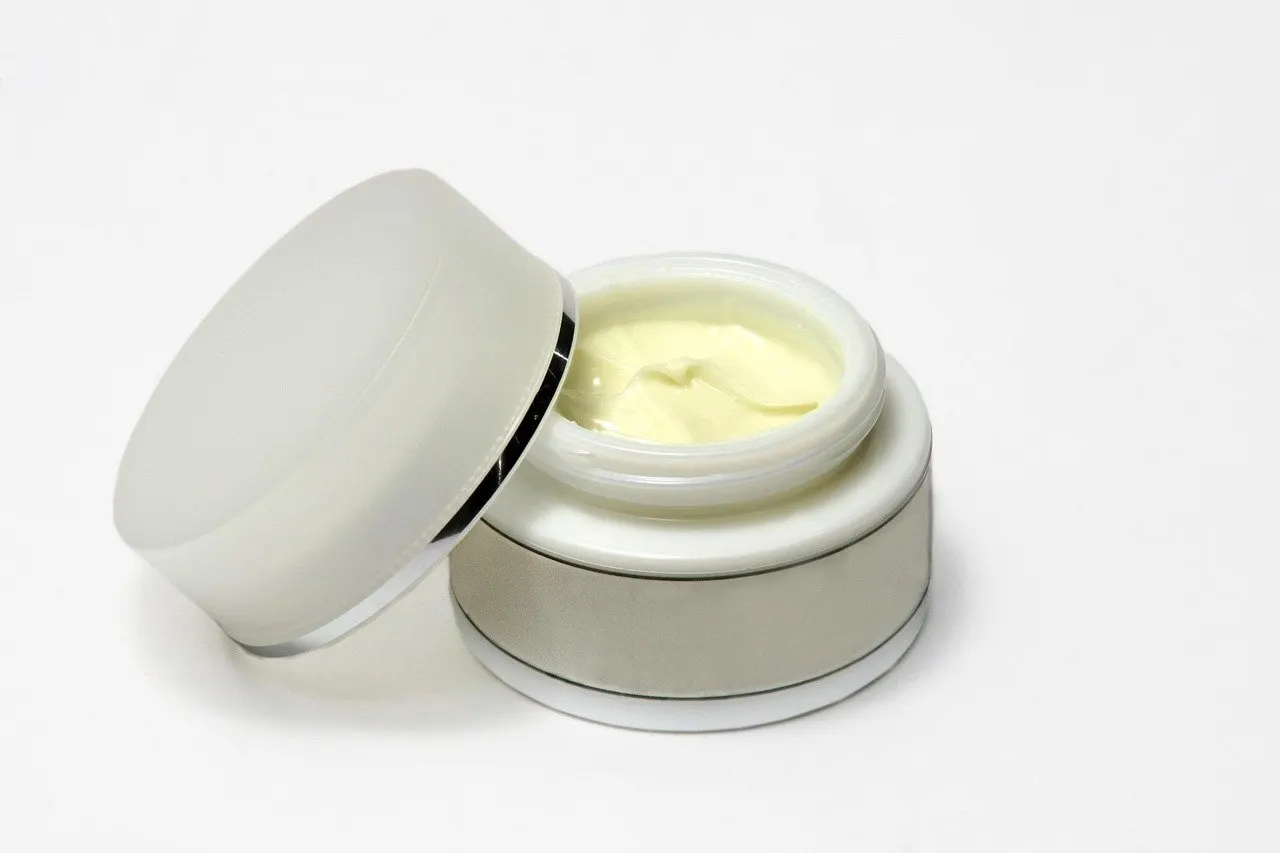 Geöffneter weißer Cremetiegel mit hellgelber Creme, der Deckel lehnt schräg daneben, heller Hintergrund ohne weitere Gegenstände
