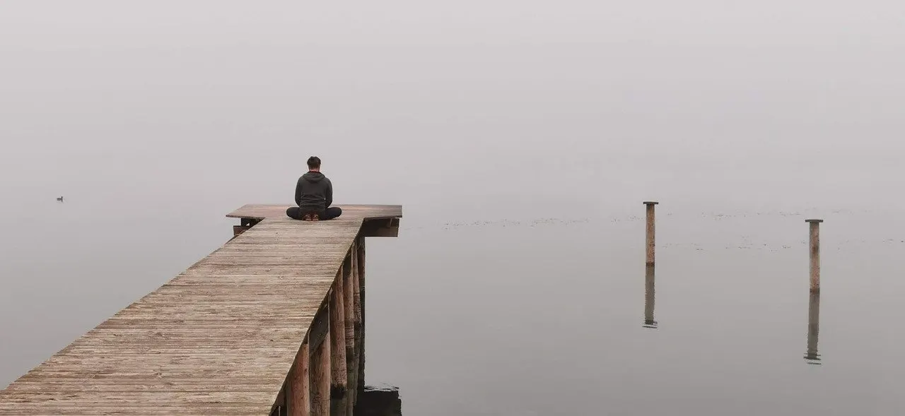 Person sitzt im Schneidersitz am Ende eines hölzernen Stegs und über einem stillen, nebligen See