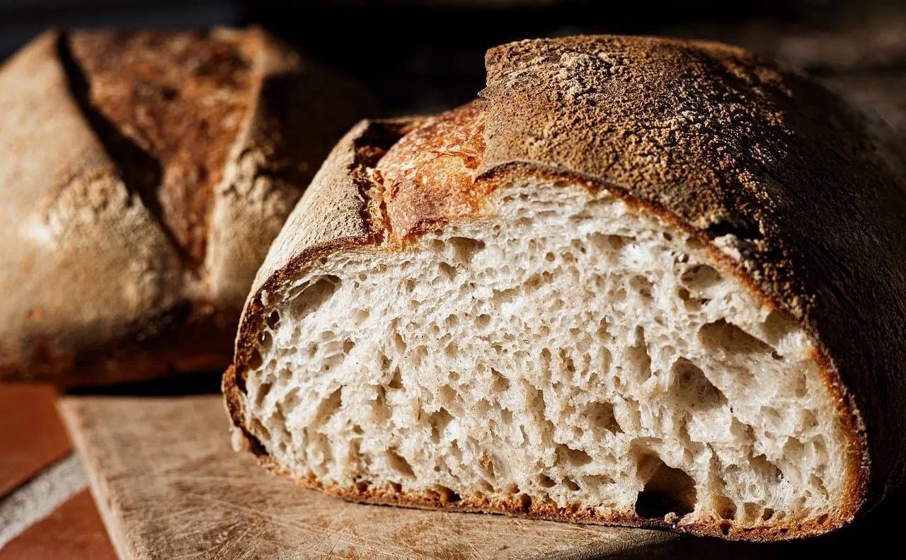 Angeschnittener Brotlaib mit grobporiger heller Krume und bemehlter dunkler Kruste auf einem Holzbrett, dahinter ein zweiter Laib