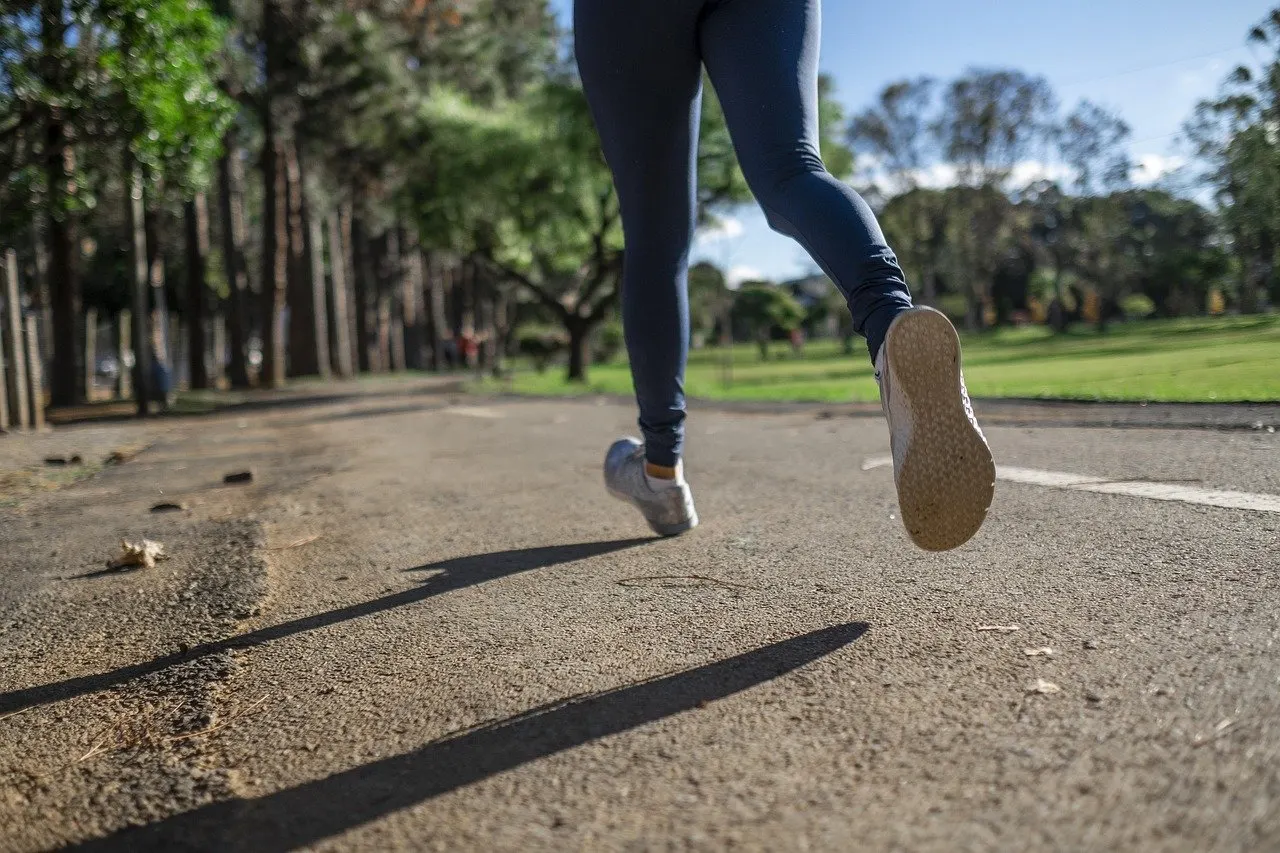 Beine einer Joggerin in dunkler Laufhose und hellen Laufschuhen auf einem asphaltierten Weg in einem sonnigen Park mit Bäumen