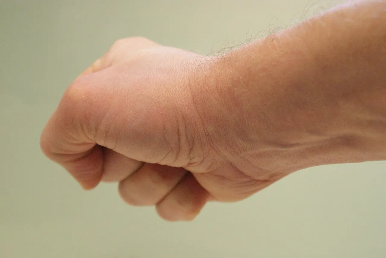 Nahaufnahme einer zur Faust geballten Hand mit Unterarm vor hellem, graugrünem Hintergrund, am Handgelenk zeichnen sich Hautfalten ab