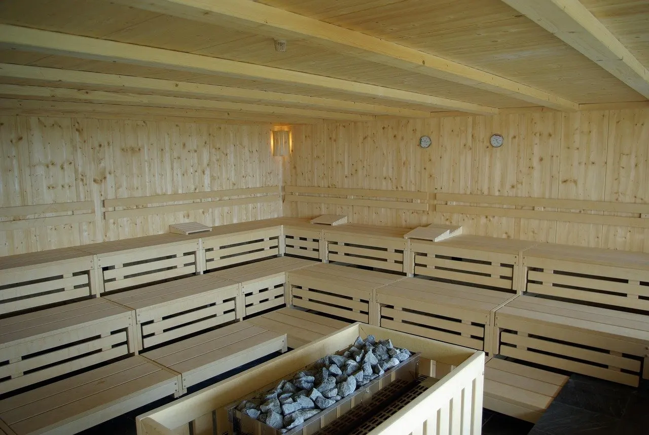Leere öffentliche Saunakabine aus hellem Holz mit mehreren Bankreihen, im Vordergrund ein Ofen mit grauen Saunasteinen