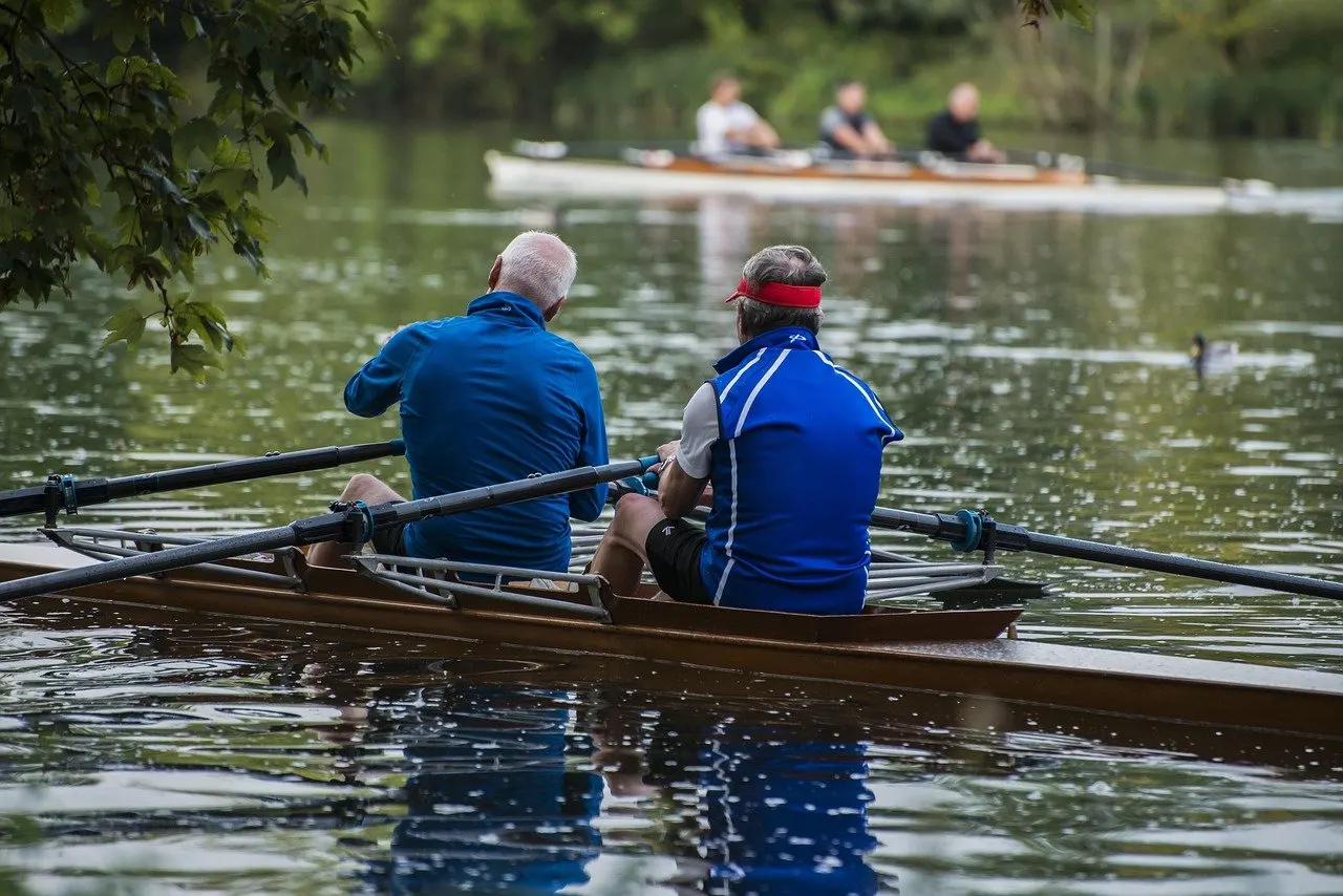 Zwei ältere Ruderer von hinten in einem Doppelzweier auf einem ruhigen Fluss, im Hintergrund ein weiteres Ruderboot und Bäume