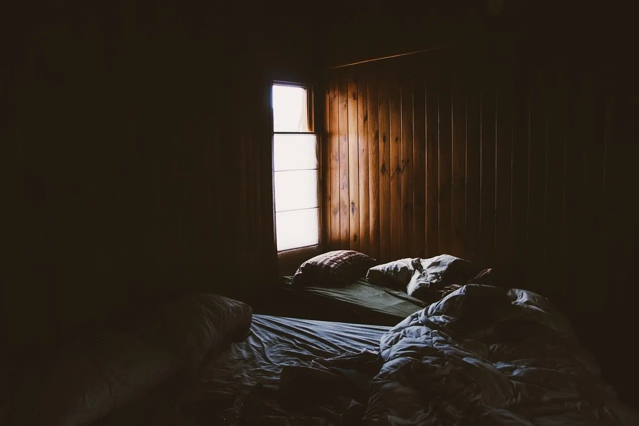 Abgedunkeltes Schlafzimmer mit Holzvertäfelung und zwei ungemachten Betten mit Decken und Kissen, Tageslicht fällt durch ein schmales Fenster