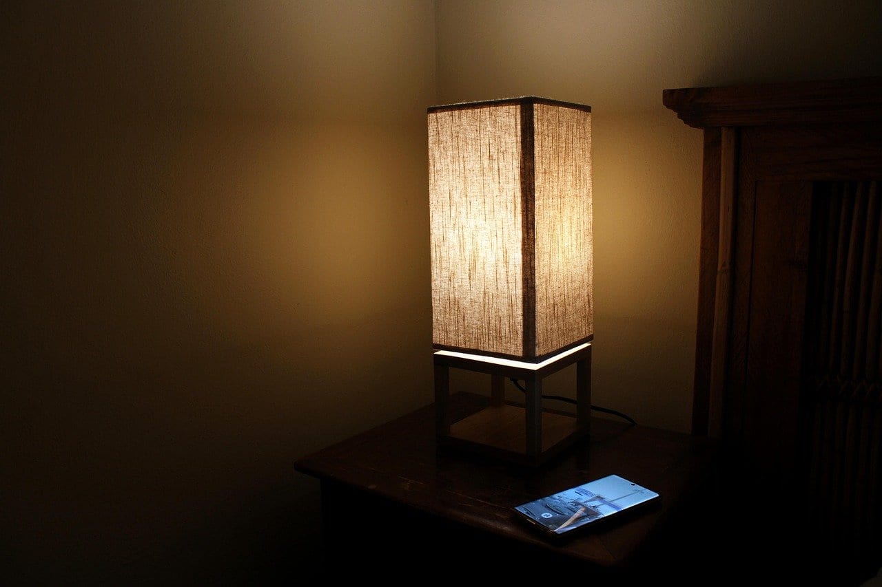 Nachttisch aus Holz mit eingeschalteter Tischlampe mit Stoffschirm und einem Smartphone mit leuchtendem Display im abgedunkelten Raum