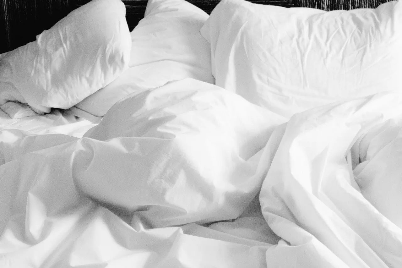 Zerwühltes Bett mit weißer Bettdecke und mehreren weißen Kopfkissen vor dunklem Kopfteil, Schwarz-Weiß-Aufnahme von schräg oben