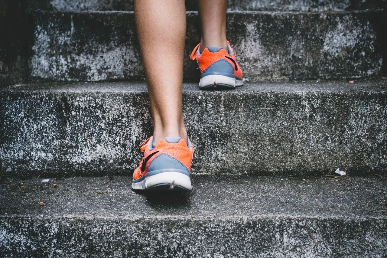 Unterschenkel einer Person in orangefarbenen Laufschuhen, die eine graue, verwitterte Betontreppe hinaufsteigt, Ansicht von hinten