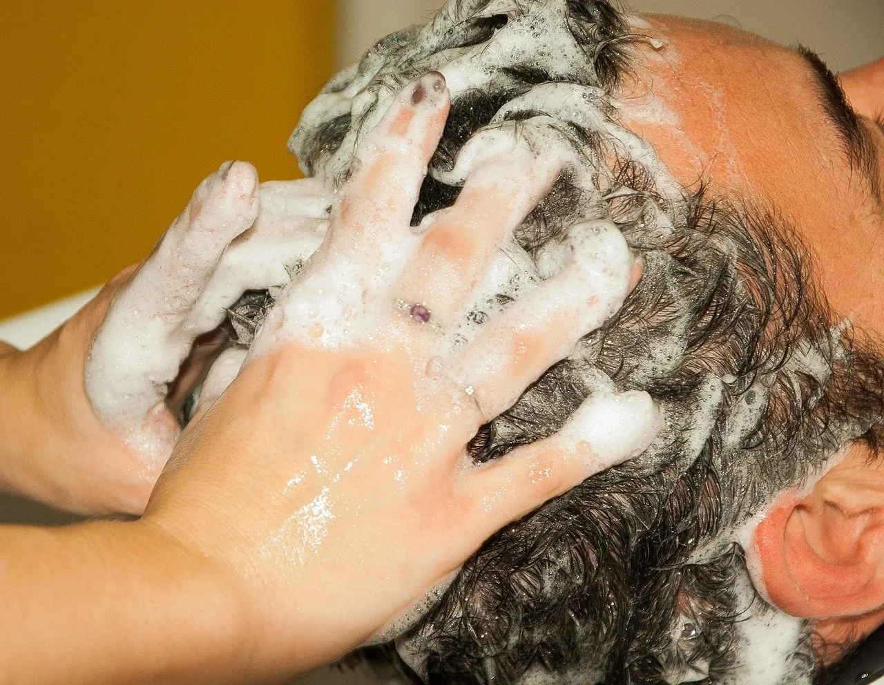 Zwei Hände massieren Shampooschaum in kurzes, dunkles nasses Haar, der Kopf ist seitlich angeschnitten, Stirn und Ohr sind sichtbar