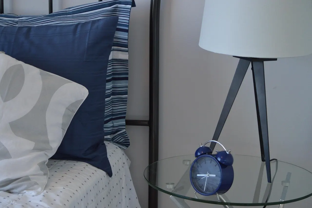 Blauer Wecker auf einem gläsernen Nachttisch neben einem Bett mit blauen und gestreiften Kissen, dahinter eine Tischlampe